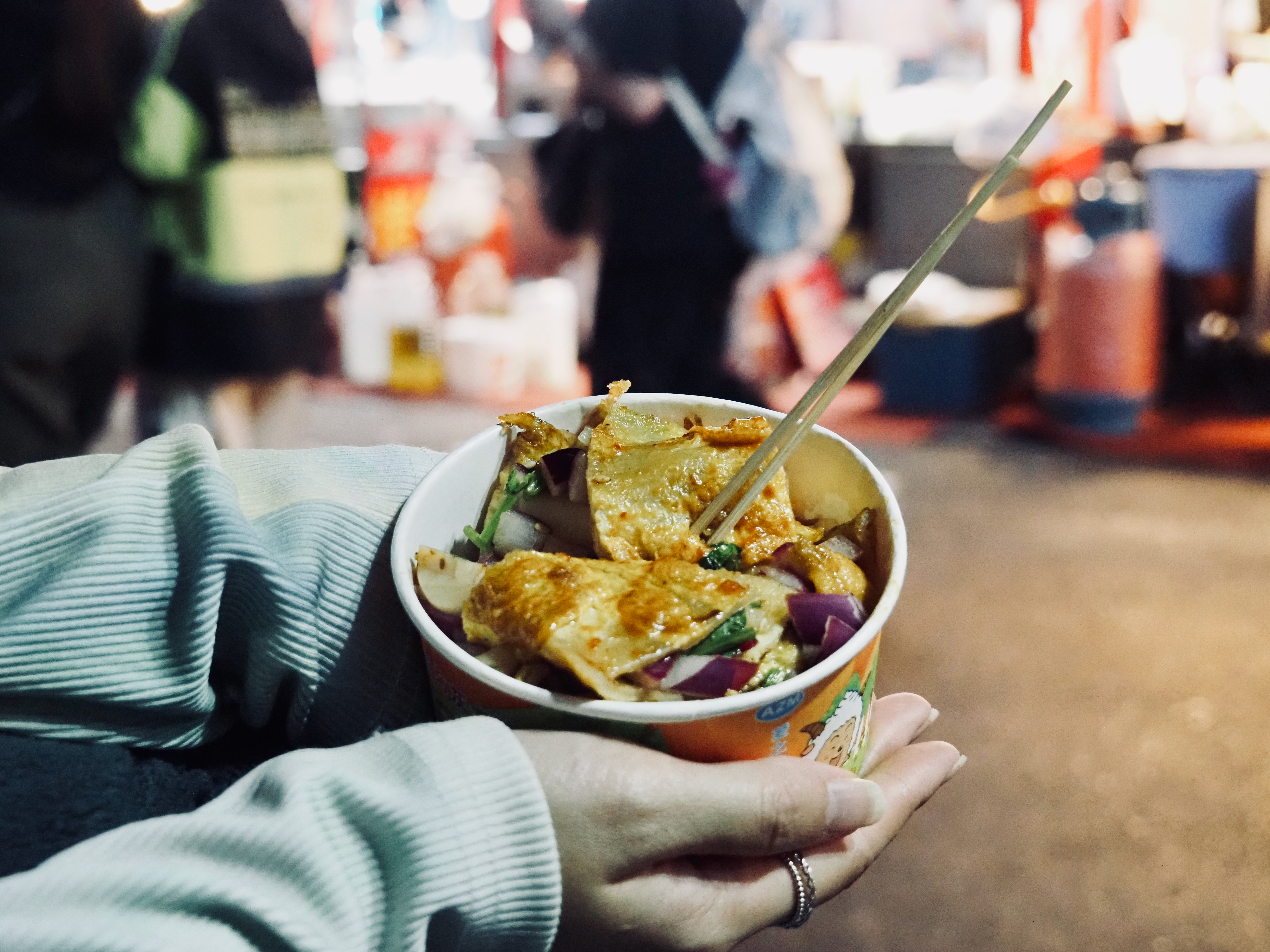 A photo of a small disposable bowl of wide noodles with veggies mixed in. In the background is a night market.