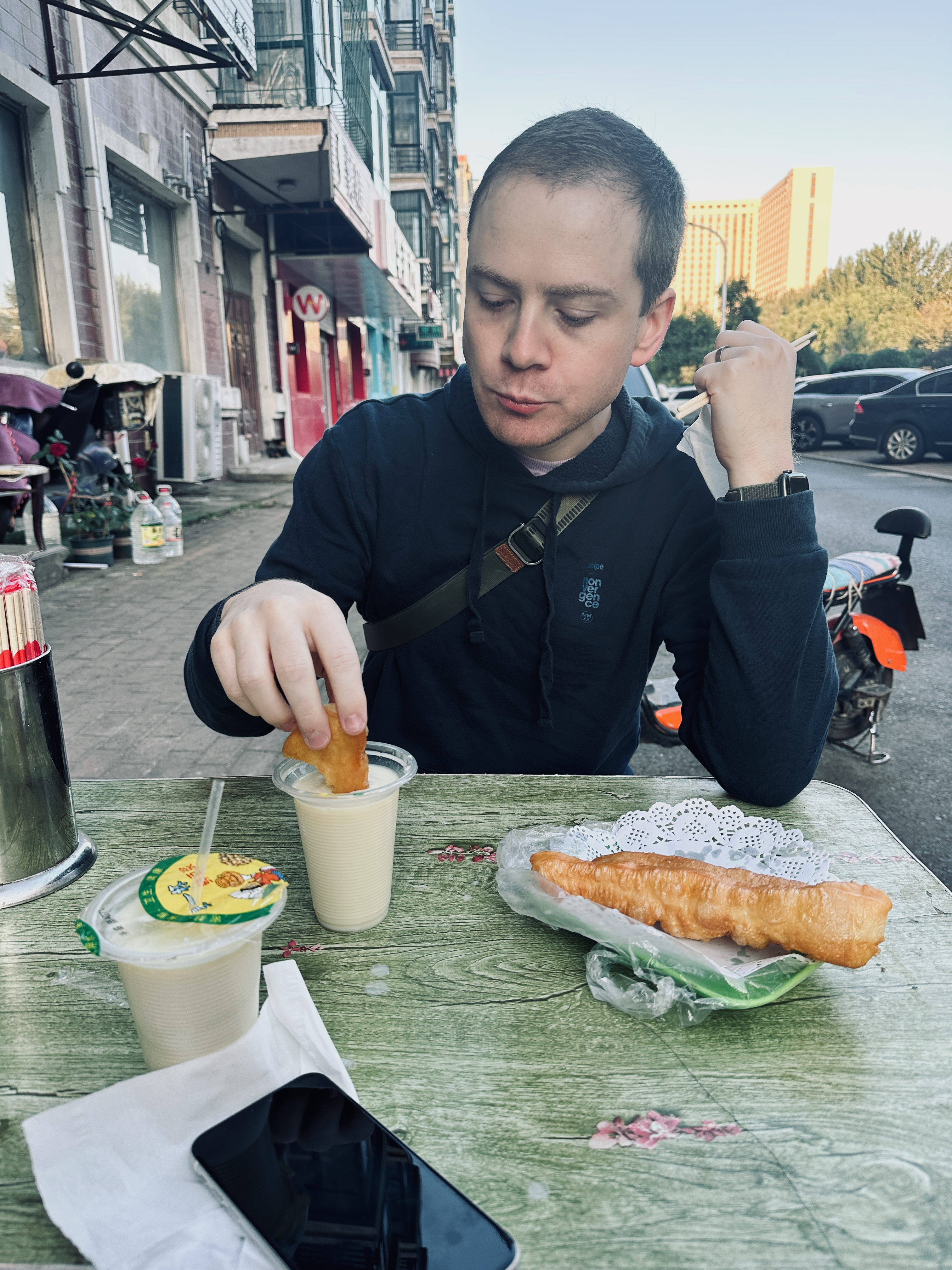 A photo of me, a white man, dipping a long donut-looking item in a cup of soy milk. We are sitting at a table on the street and the sun is rising.